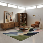 A modern mid-century styled living room featuring a large abstract geometric area rug in shades of green, blue, pink, beige, and black on a terrazzo floor. The room includes a wooden sideboard with a framed abstract print, a tall wooden bookshelf, a brown leather armchair, a gold floor lamp, and a matching wooden coffee table.