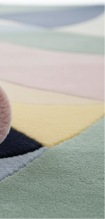 Close-up of a soft, plush area rug with a pastel geometric pattern, showing textures and colors of pink, yellow, sage green, navy, and light blue.