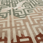 A close-up of a textured area rug featuring a geometric maze pattern in sage green, cream, and beige, with a white bird motif in the upper left and terracotta accents near the bottom.
