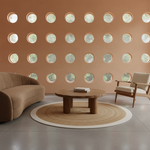 A modern living room scene with a curved tan sofa, a round wooden coffee table, a cream armchair, and a woven round rug in front of a terracotta-colored wall with evenly spaced circular windows.
