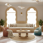 A beautifully styled bohemian living room featuring a cream-colored sofa, a pink armchair with a beige throw blanket, a teal gradient accent chair, a round woven jute rug, and a central wooden coffee table. The room is decorated with two large arched windows, hanging pendant lights, and potted plants.