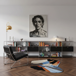 A large black and white portrait painting of a woman hangs on a light gray wall above a long metal bookshelf in a modern living room. A black leather chair with a chrome frame sits to the left, and a colorful abstract area rug lies on the wood floor.
