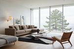A modern living room featuring a taupe sectional sofa, a black and white geometric area rug, a wooden armchair with a white cushion, and a glass coffee table. Large floor-to-ceiling windows offer a view of a pine tree, set against a clean white wall.